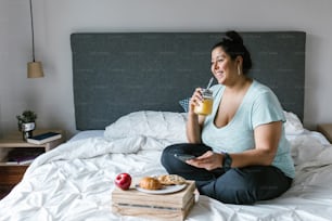 Ragazza latina formosa con smartphone che fa colazione e beve succo d'arancia a letto in america latina, donna plus size