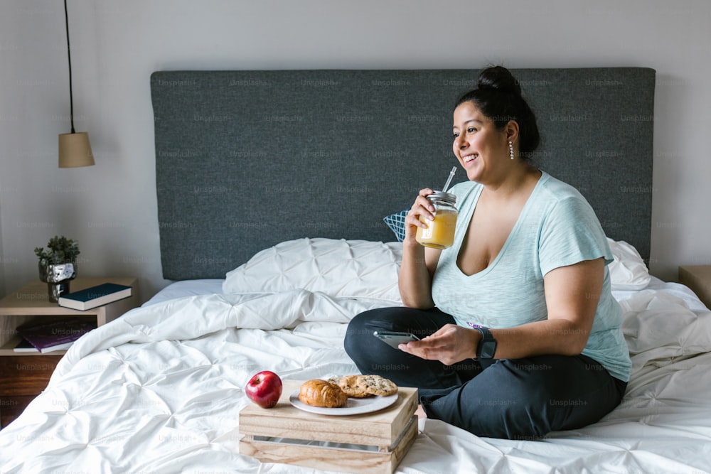 Curvy latin girl with smartphone having breakfast and drinking orange juice in bed in Latin America, plus size woman