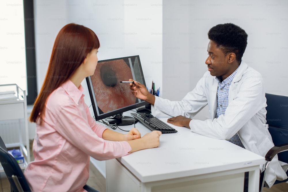 Confident African American male doctor in his modern office, explaining results of endoscopy to young female Caucasian patient, pointing on the image of digestive tract on computer