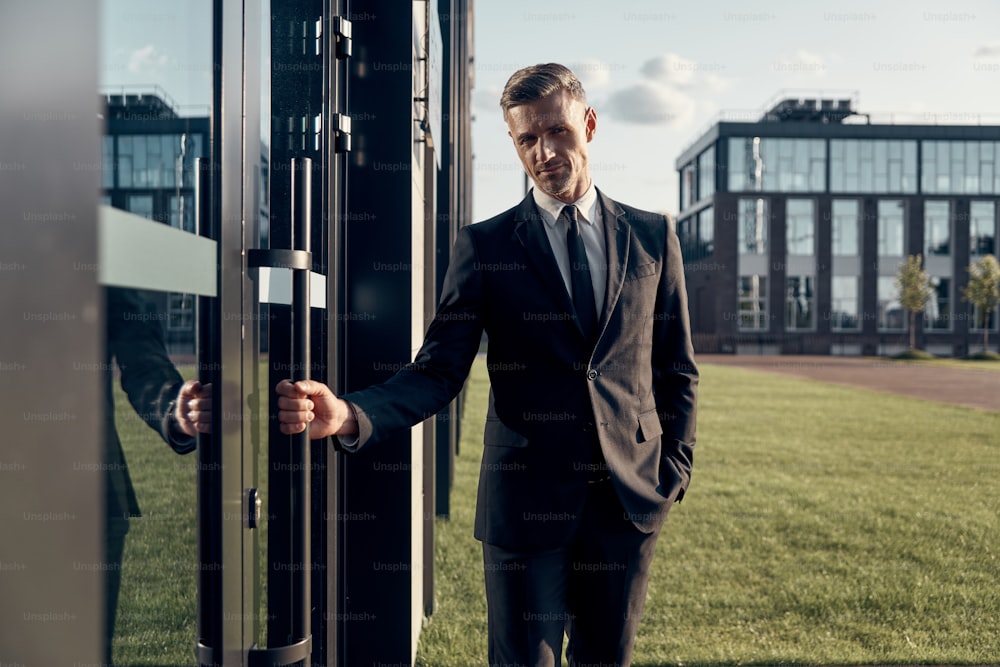 Confident mature businessman opening the door of business centre and looking at camera
