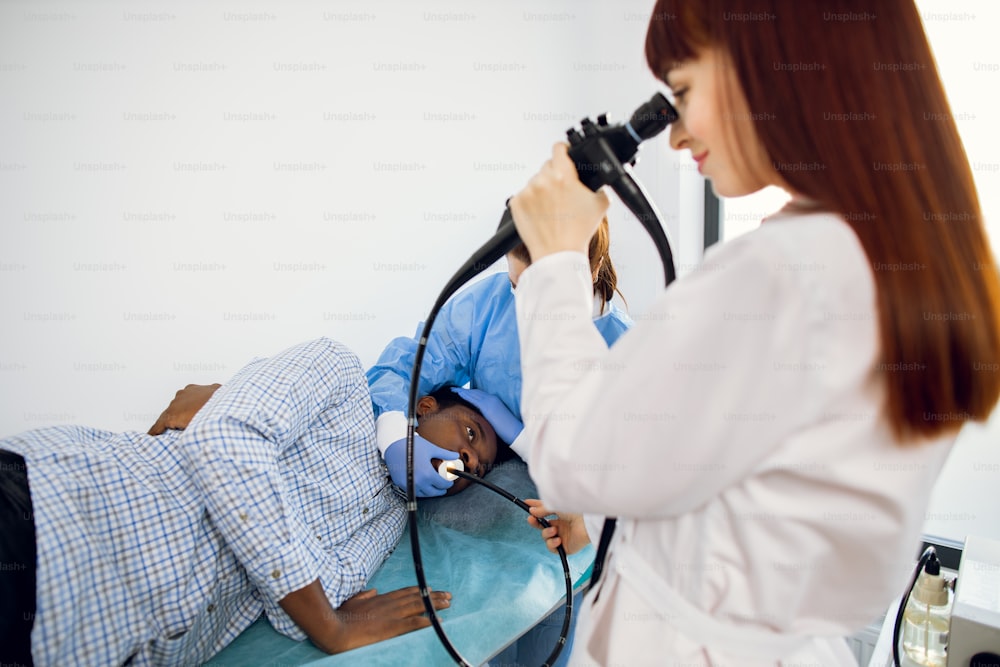 病院のソファに横たわる若いアフリカ系アメリカ人男性患者と、2人の白人女性医師のチームが胃十二指腸内視鏡検査の処置を行っている