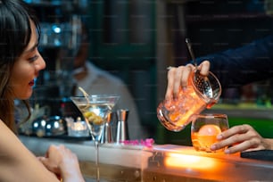 Mujer asiática de confianza sentada en la barra del bar bebiendo bebidas alcohólicas y hablando con el barman en el club nocturno. Camarero mixólogo masculino preparando bebidas alcohólicas para el cliente. Concepto de bar de pequeñas empresas y vida nocturna de la ciudad