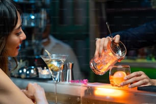 Confidence Asian woman sitting at bar counter drinking alcoholic drink and talking to barman in nightclub. Male mixologist bartender preparing alcohol drink to customer. Small business bar and city nightlife concept