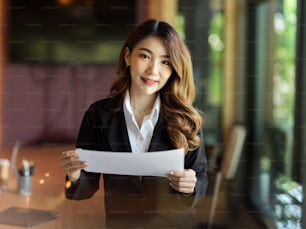 Business executive female holding paper bill, letter, business report, standing in office board room, portrait