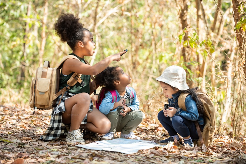 Group of Diversity little girl friends with backpack hiking together at forest mountain in summer sunny day. Three kids having fun outdoor activity sitting and looking at the map exploring the forest.