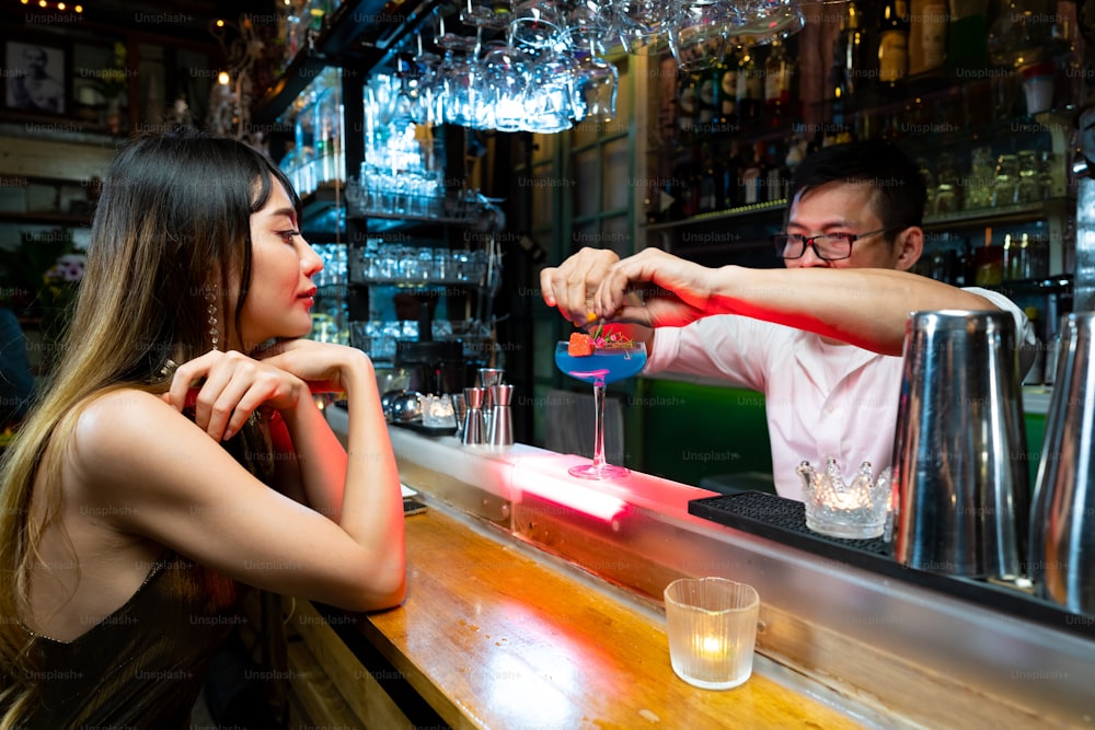 Confiance Femme asiatique assise au comptoir du bar buvant une boisson alcoolisée et parlant au barman dans une boîte de nuit. Mixologue barman préparant une boisson alcoolisée au client. Concept de bar pour petites entreprises et de vie nocturne urbaine