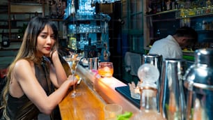 Confidence Asian woman sitting at bar counter drinking alcoholic drink and talking to barman in nightclub. Male mixologist bartender preparing alcohol drink to customer. Small business bar and city nightlife concept