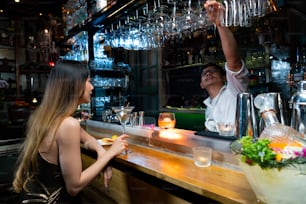 Confidence Asian woman sitting at bar counter drinking alcoholic drink and talking to barman in nightclub. Male mixologist bartender preparing alcohol drink to customer. Small business bar and city nightlife concept