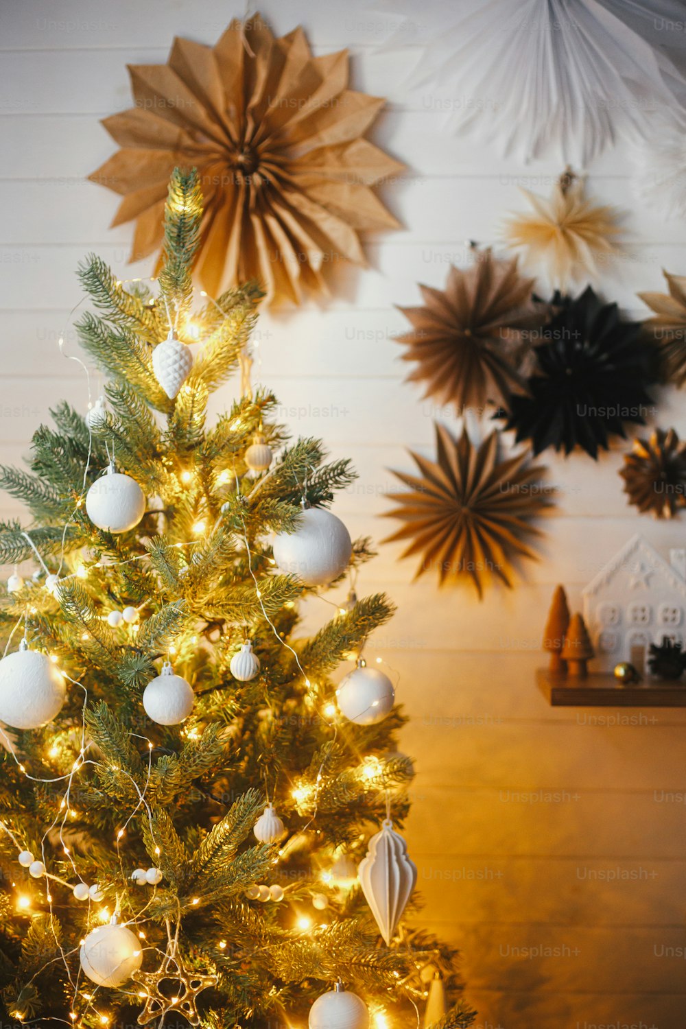 Foto Elegante árbol de Navidad decorado con modernos adornos blancos y luces  doradas sobre el fondo de una pared blanca con estrellas de papel.  Habitación escandinava festiva atmosférica. Nochebuena – Árbol de