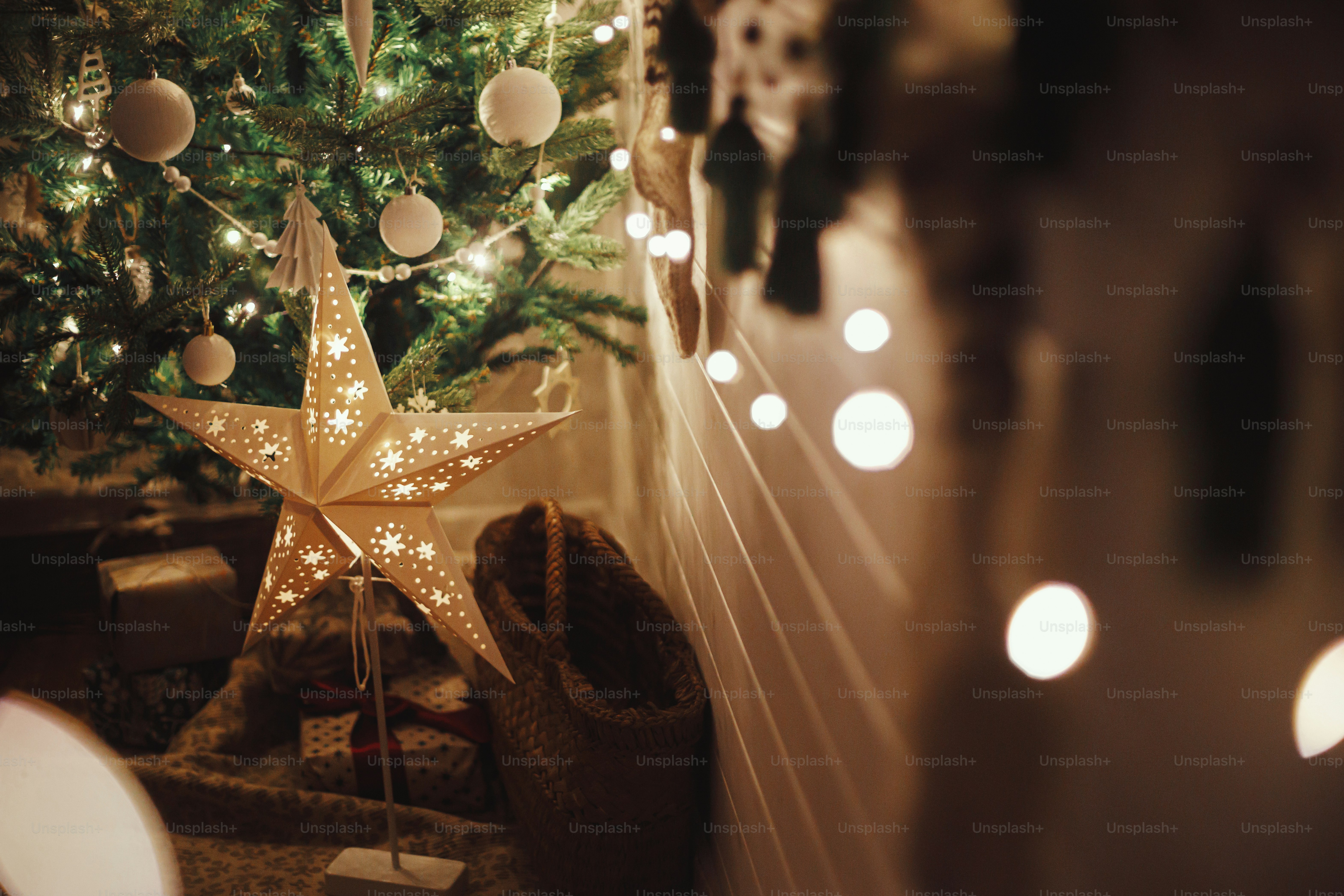 Stylish Christmas star, tree with white baubles, boho ornaments, golden lights and gifts in atmospheric evening room. Festive scandinavian room at eve. Magic time. Space for text. Merry Christmas!