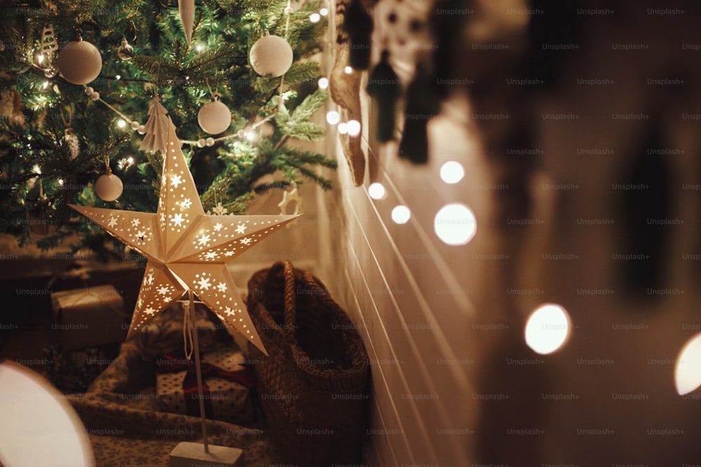 Elegante estrella de Navidad, árbol con adornos blancos, adornos boho, luces doradas y regalos en la sala de noche atmosférica. Sala festiva escandinava en la víspera. Tiempo mágico. Espacio para el texto. ¡Feliz Navidad!
