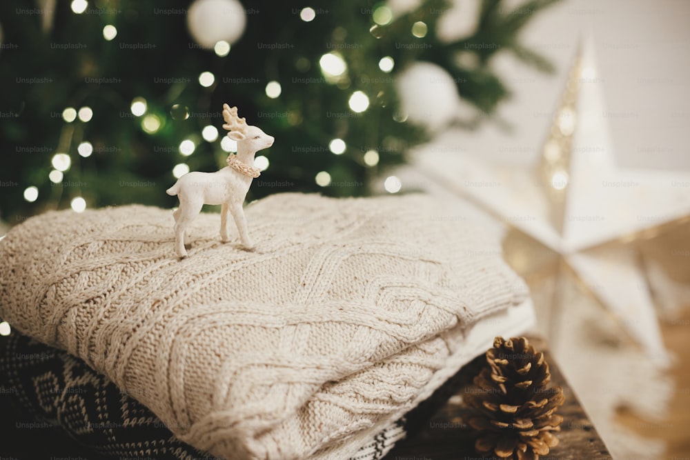 Cozy knitted sweaters with reindeer toy and pine cone on rustic wood on background of christmas tree lights. Pile of stylish sweaters in festive decorated scandinavian room. Atmospheric winter time