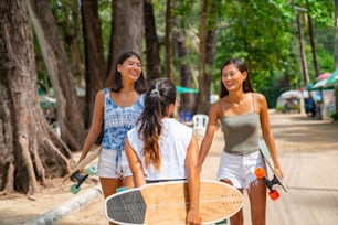 Group of Happy Asian woman girl friends carry skateboard walking with talking together by the beach. Female friendship enjoy summer outdoor activity lifestyle play extreme sport surf skate together