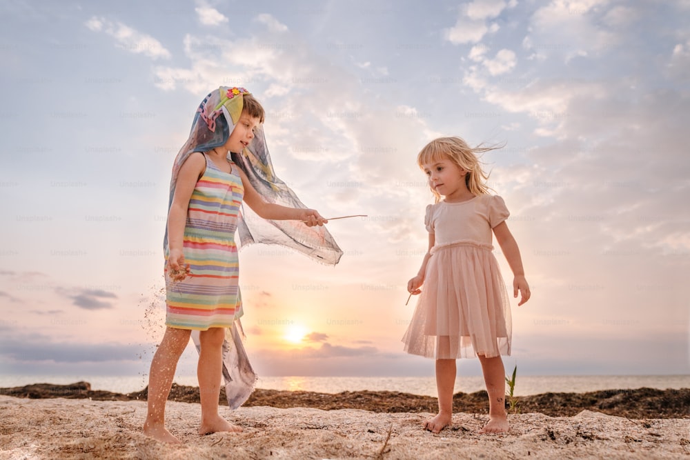 Two little girls having fun time playing fighting with wands on the beach at summer sunset