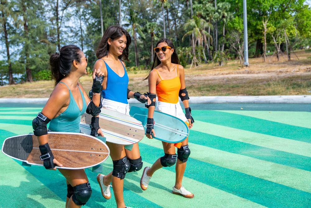 幸せなアジアの女性の女の子の友達のグループは、スケートボードパークで一緒に話しながらスケートボードを運びます。女性の友情は、アウトドアアクティビティのライフスタイルを楽しみ、エクストリームスポーツのサーフスケートを一緒にプレイします