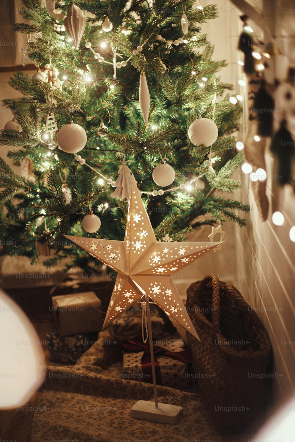 Stylish Christmas star, tree with white baubles, boho ornaments, golden lights and gifts in atmospheric evening room. Festive scandinavian room at eve. Magic time. Space for text. Merry Christmas!