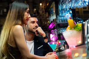 バーカウンターに座ってアルコール飲料を飲み、ナイトクラブでバーテンダーと話している自信のあるアジア人女性。お客さんにお酒を振る舞う男性ミクソロジストのバーテンダー。スモールビジネスバーと街のナイトライフのコンセプト