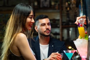 Confidence Asian woman sitting at bar counter drinking alcoholic drink and talking to barman in nightclub. Male mixologist bartender preparing alcohol drink to customer. Small business bar and city nightlife concept