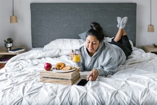 Chubby hispanic woman having breakfast in bed while using the phone in Latin America, plus size female