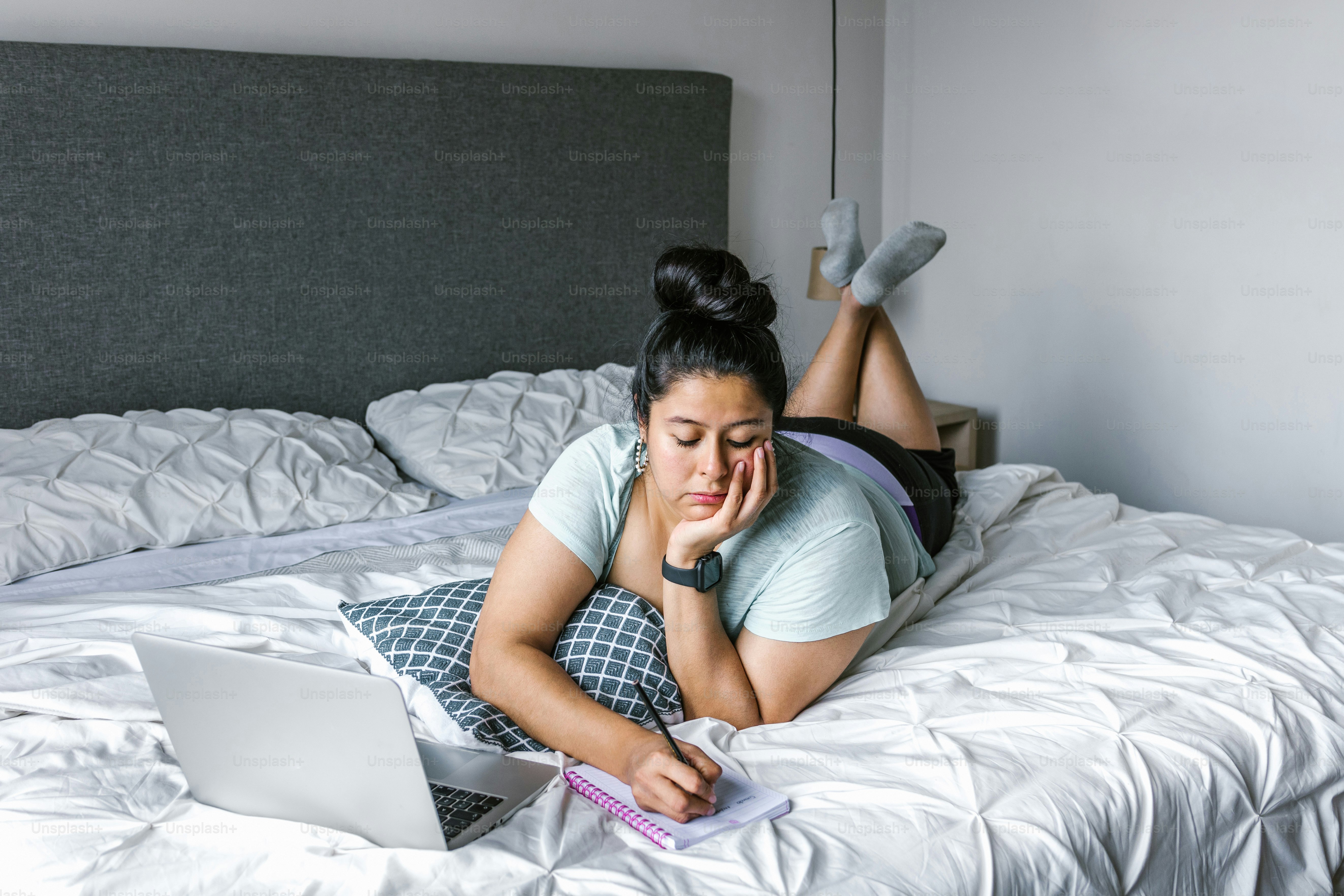 Wanita muda Latin ukuran plus berbaring di tempat tidur membuat catatan di buku catatan menggunakan komputer di Amerika Latin