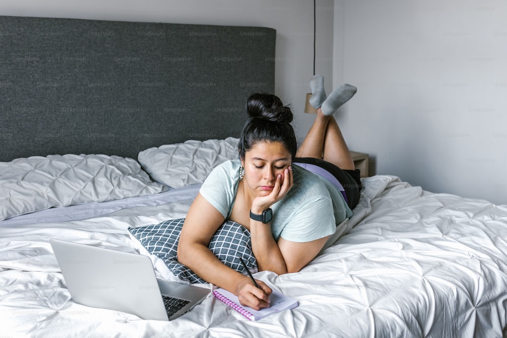 Giovane donna latina plus size sdraiata a letto che prende appunti in taccuino usando il computer in America Latina