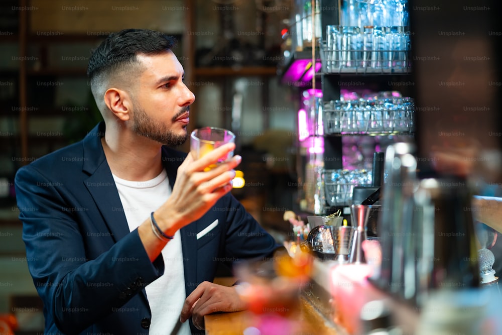 Vertrauen kaukasischer Mann, der an der Bartheke sitzt, genießt es, auf alkoholische Getränke zu warten und mit Barkeeper im Nachtclub zu sprechen. Männlicher Barkeeper bereitet Alkoholgetränk im Shaker mit Eisball auf Steinglas zu.