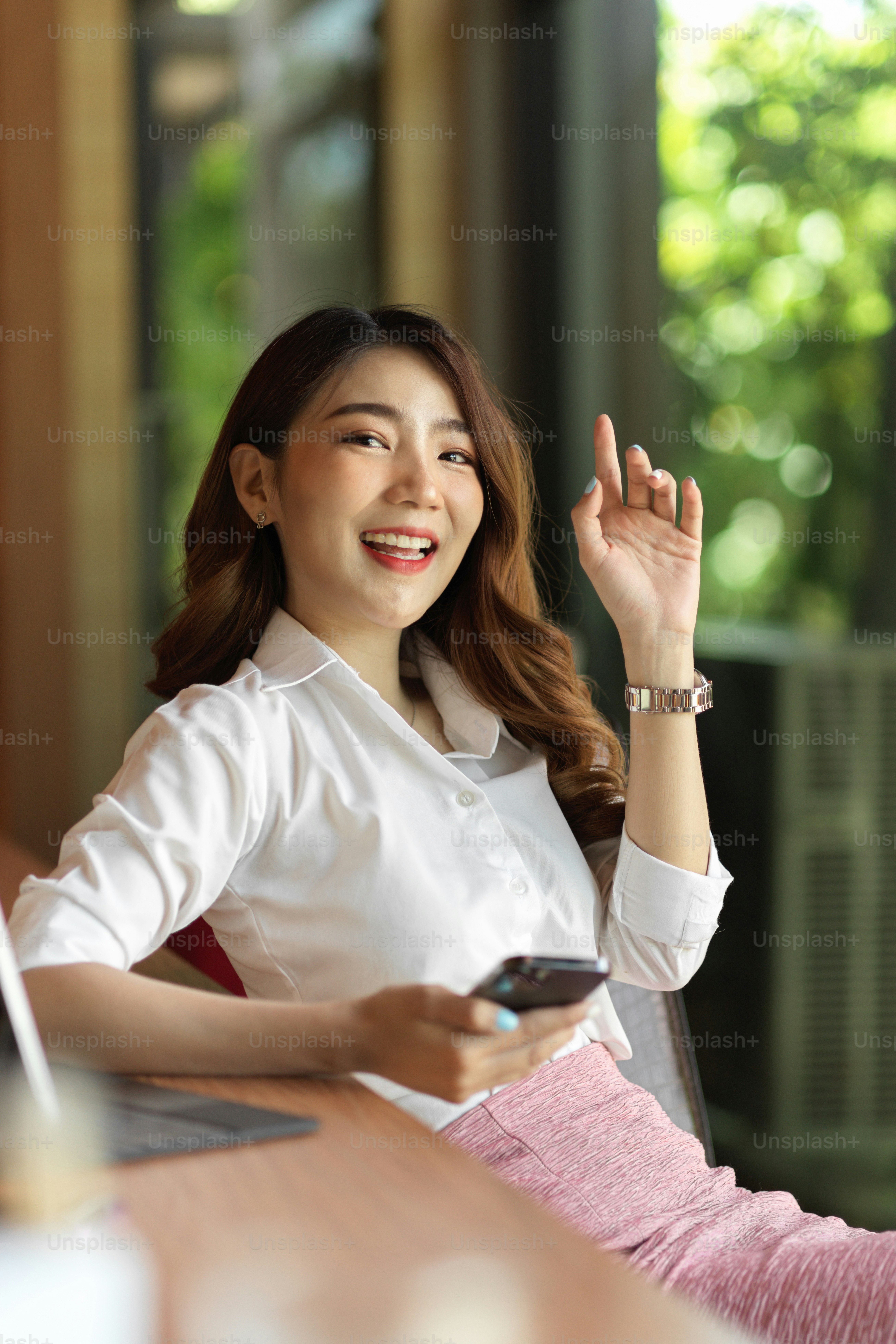 Portrait of attractive friendly asian businesswoman raise hand to greeting her co-worker, phone in another hand, having question while meeting