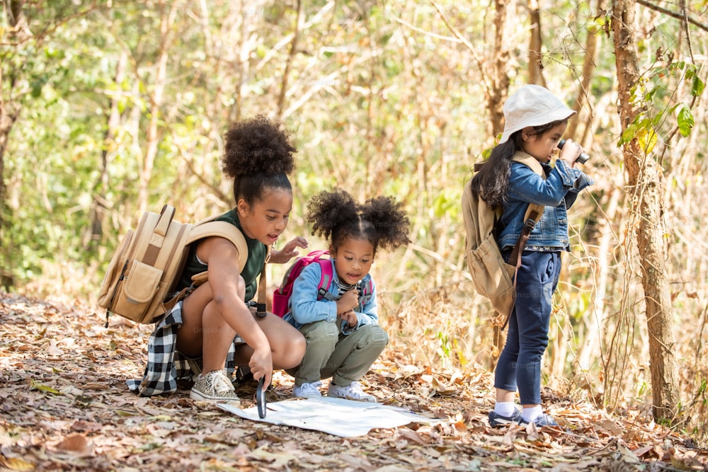 Gruppe der Vielfalt kleine Freundinnen mit Rucksack wandern gemeinsam am Waldberg im Sommer sonnigen Tag. Drei Kinder, die Spaß an Outdoor-Aktivitäten haben, sitzen und schauen auf die Karte, um den Wald zu erkunden.