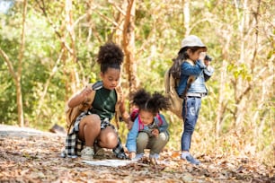 Group of Diversity little girl friends with backpack hiking together at forest mountain in summer sunny day. Three kids having fun outdoor activity sitting and looking at the map exploring the forest.