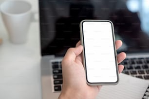 Image of a businessman holding smartphone a blank screen, laptop placed on an office table. Mock up.