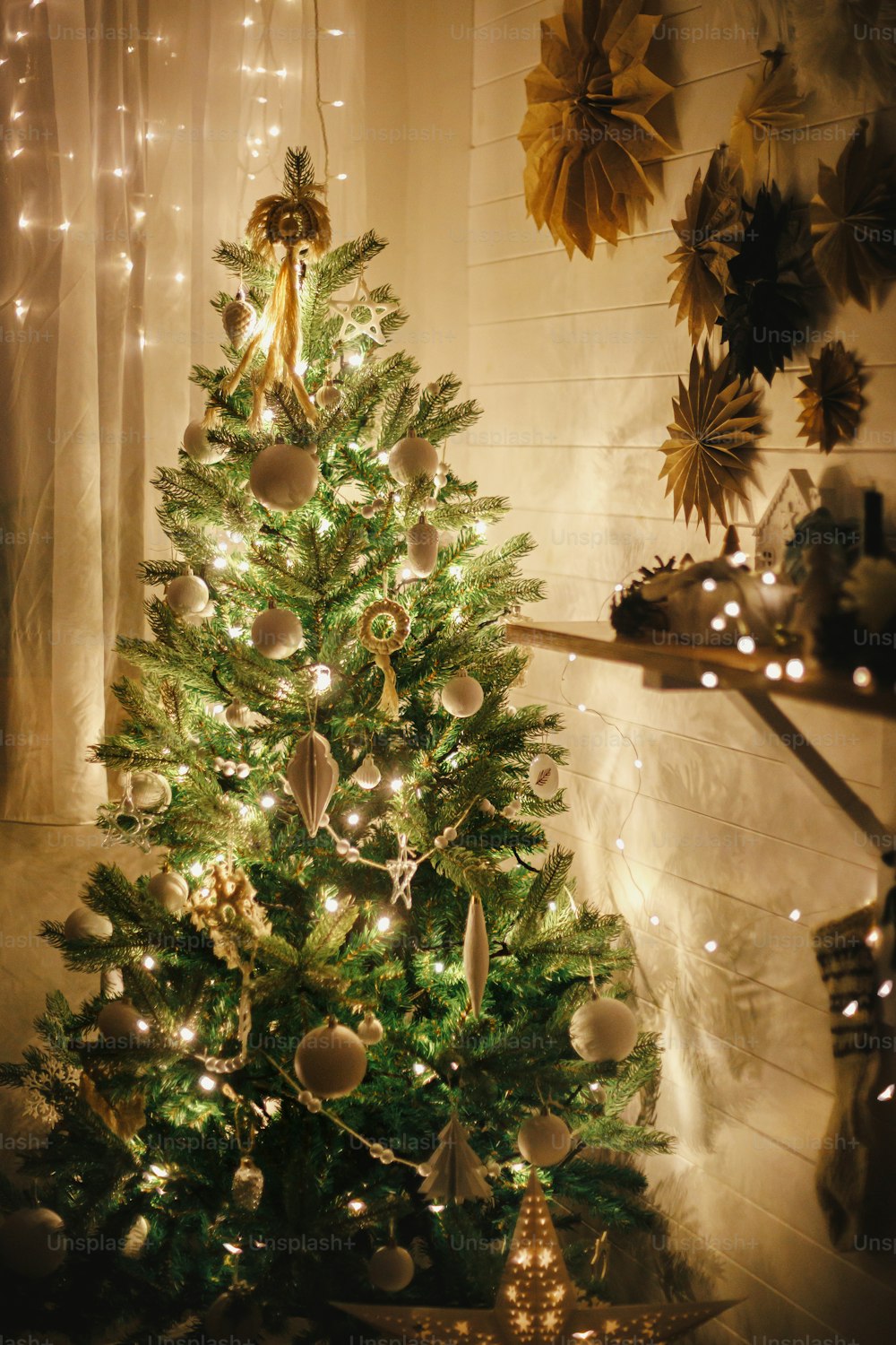 Stilvoller Weihnachtsbaum geschmückt mit modernen weißen Kugeln, Boho-Ornamenten und goldenen Lichtern auf dem Hintergrund von Papiersternen an Wand und Strumpf. Stimmungsvolles festliches skandinavisches Zimmer.  Magische Zeit