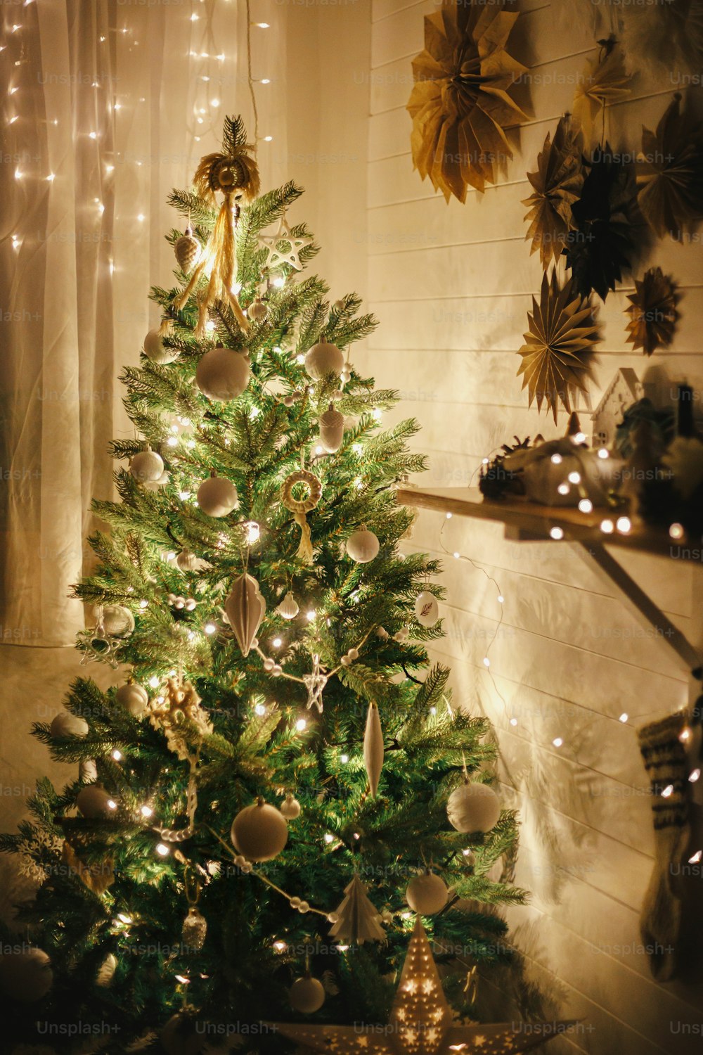 Stylish Christmas tree decorated with modern white baubles, boho ornaments and golden lights on background of paper stars on wall and stocking. Atmospheric festive scandinavian room.  Magic time