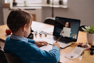 Un niño preadolescente usa una computadora portátil para hacer una videollamada con su maestra. La pantalla muestra una conferencia en línea con un profesor explicando el tema de la clase.