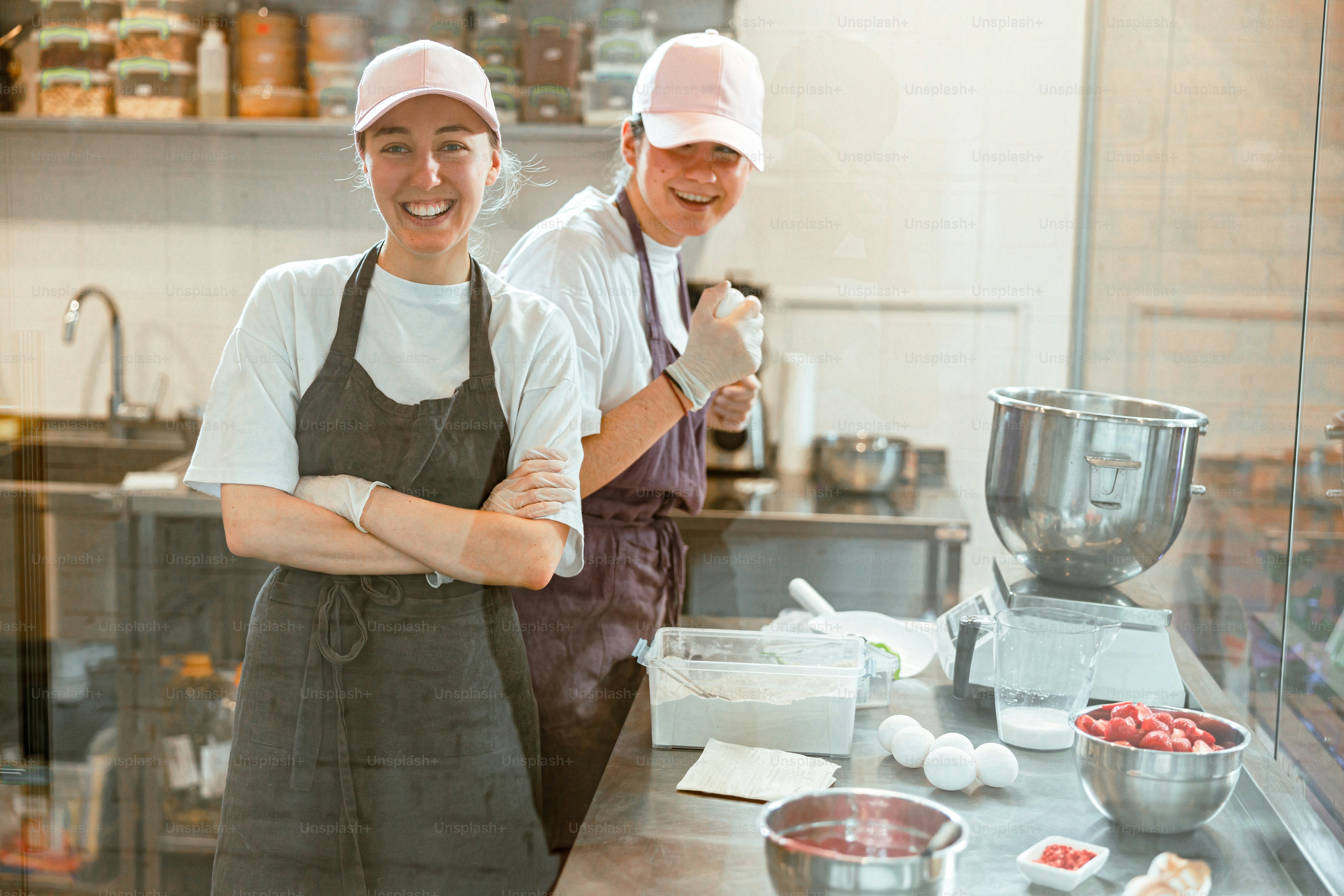 Positive women colleaguesin aprons smile to camera standing near metal counter with food products for dough in craft bakery shop