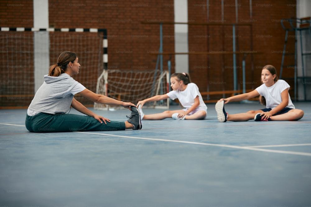 Sportlehrerin und ihre Schüler wärmen sich auf und strecken ihre Beine in einer Klasse in der Grundschulturnhalle.