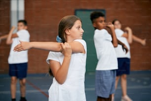 학교 체육관에서 체육 교육을 하는 동안 워밍업하는 초등학생 그룹. 초점은 팔을 뻗는 소녀에게 있습니다.