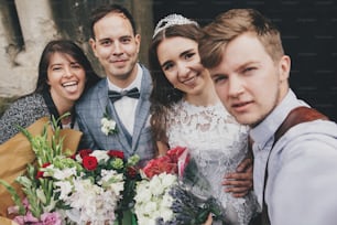 Stylish happy bride and groom taking selfie with family and smiling on background of church after holy matrimony. Beautiful emotional wedding couple with guests making selfie photo