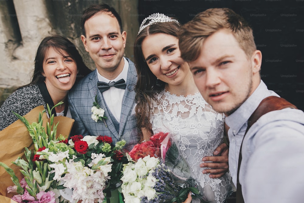 Stylish happy bride and groom taking selfie with family and smiling on background of church after holy matrimony. Beautiful emotional wedding couple with guests making selfie photo