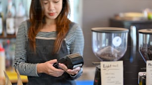 Asian barista using payment terminal, entering drinking cost for customer payment at coffee shop, contactless technology in hospitality service