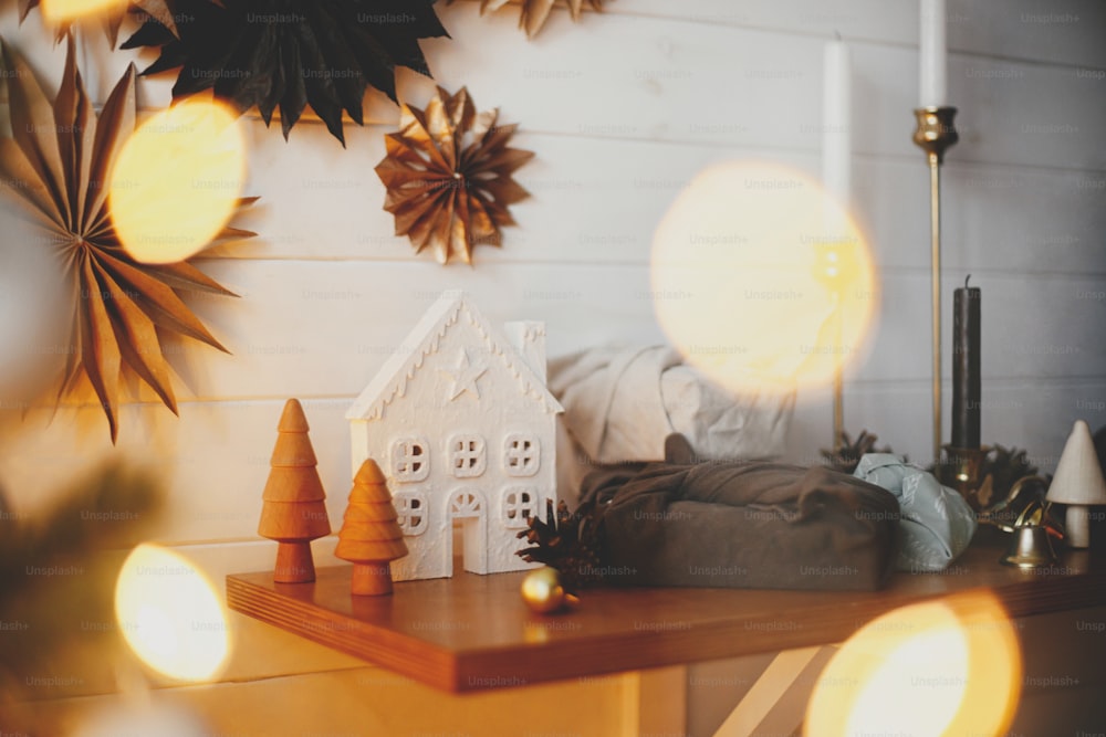 Stylish little christmas trees, house, plastic free gifts on background of white wooden wall with paper stars and golden lights bokeh. Space for text. Festive decorated scandinavian room