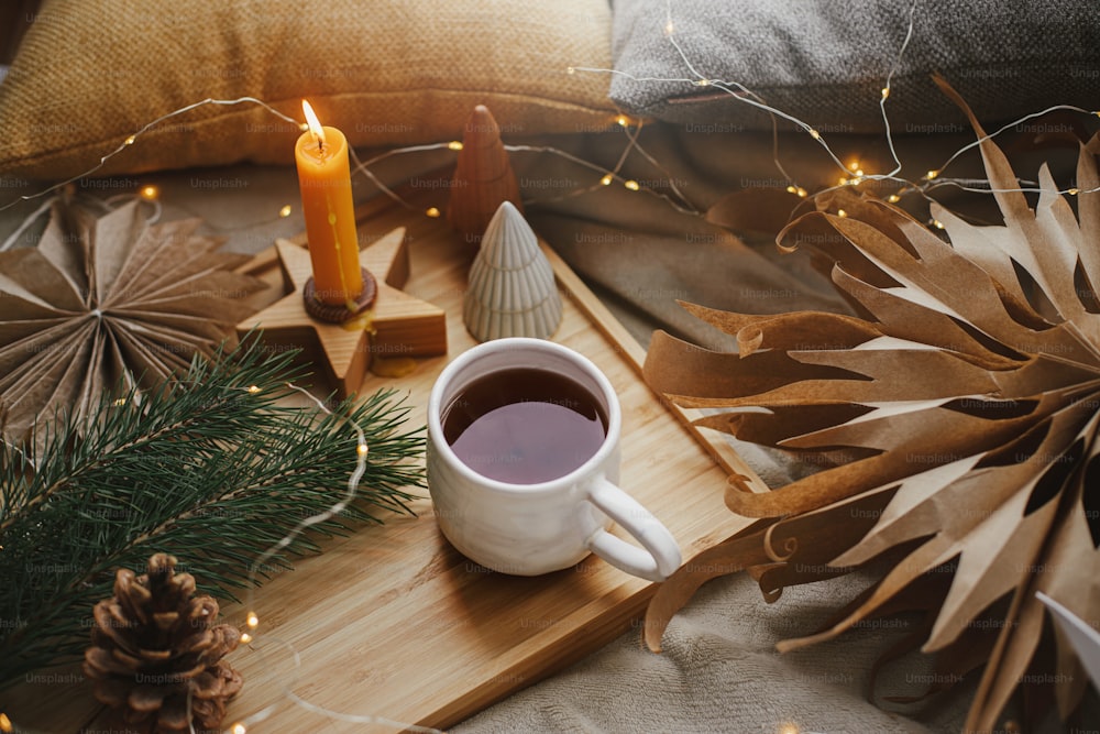 Warm cup of tea on soft bed with christmas stars, golden lights, pine trees, candle and pillows in scandinavian room. Cozy home. Atmospheric magic moment. Hygge winter holidays