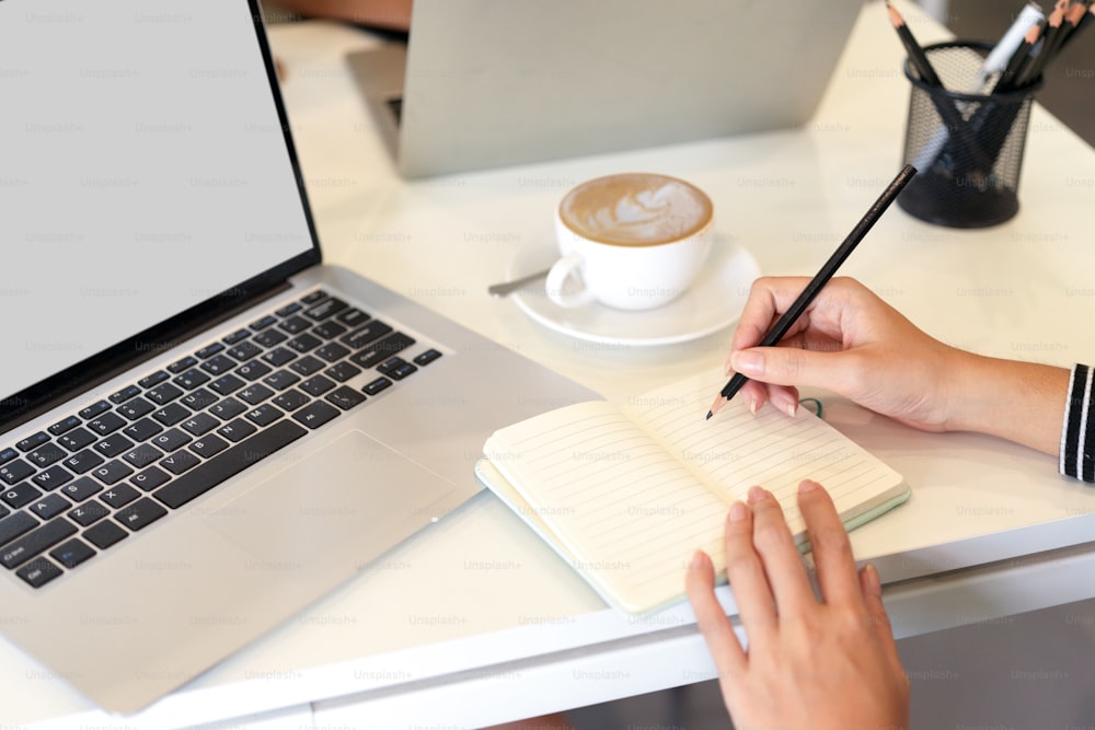 Cropped image of woman take notes on notebook next to laptop computer mockup and coffee mug at office