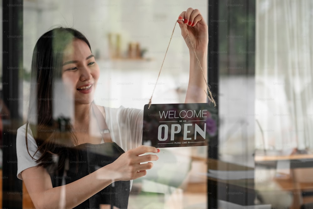 Retrato de uma jovem barista sorridente no avental segurando a placa aberta enquanto estava em pé em seu café. elegante café asiático pessoal feminino virar placa de porta de manhã na loja própria pequena empresa.