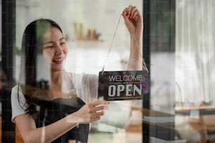 Retrato de una joven barista sonriente en delantal sosteniendo un letrero abierto mientras está de pie en su café. El personal femenino de la elegante cafetería asiática gira la placa de la puerta por la mañana en la pequeña empresa de la tienda propia.