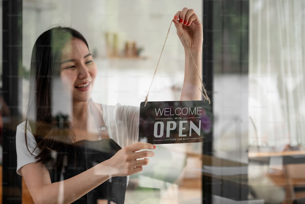 Portrait of smiling young barista girl in apron holding open sign board while standing at her cafe. elegant asian coffee shop female staff turn door plate in the morning in own store small business.