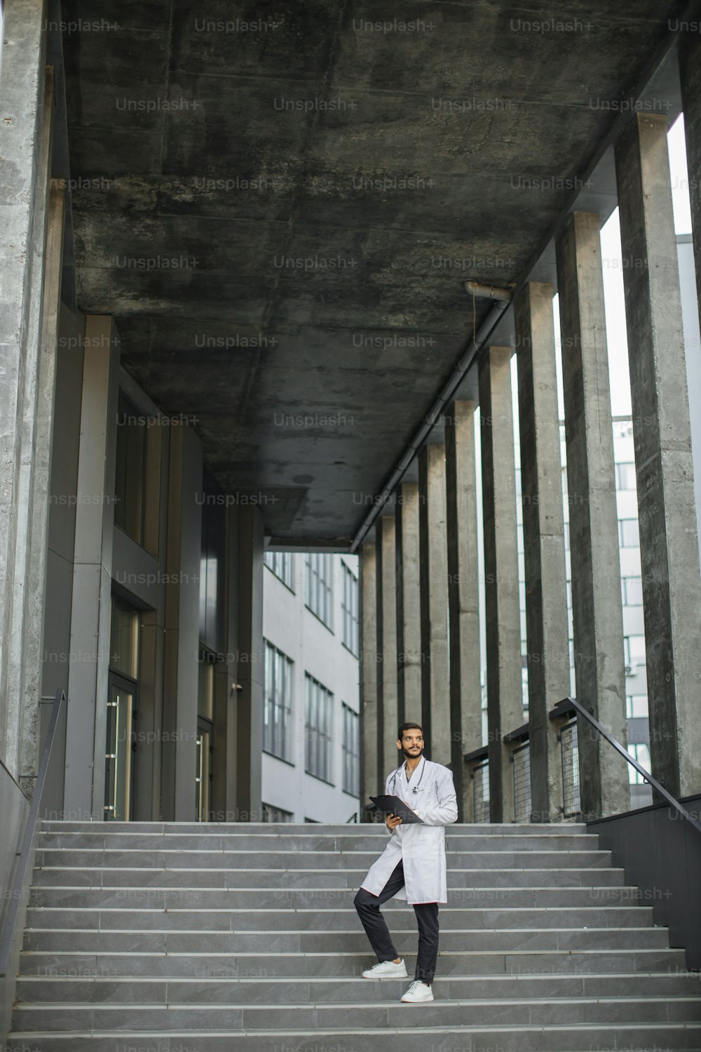 Figura intera di giovane lavoratore sanitario arabo esperto e bello, che prende appunti sugli appunti della cartella, in piedi sulle scale di un edificio moderno. Medico indiano professionista che compila la cartella clinica all'aperto