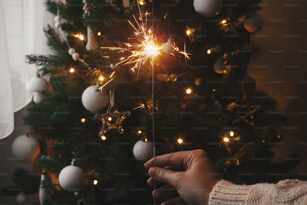 Happy New Year! Hand holding burning sparkler on background of christmas tree lights in festive room. Firework bengal glowing in woman hand. Space for text. Atmospheric moment. Merry christmas