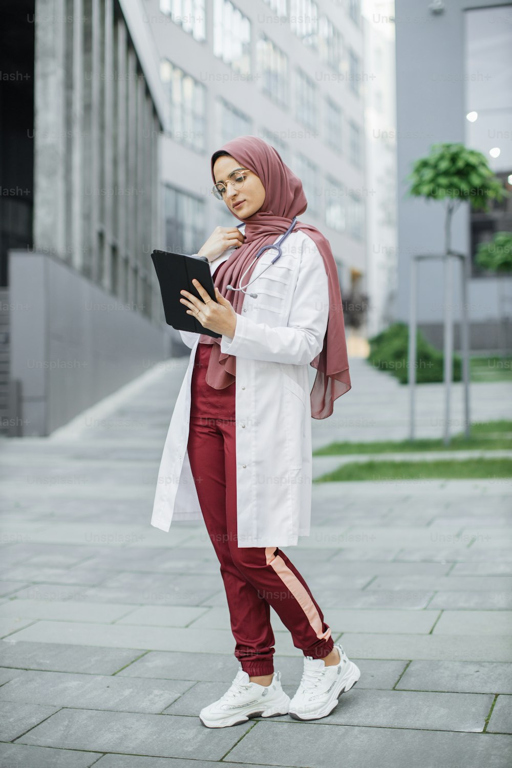 Muslimische Ärztin im Hijab, trägt Peelings und weißen Kittel, arbeitet mit Patientenakten auf digitalem Tablet vor der modernen Klinik