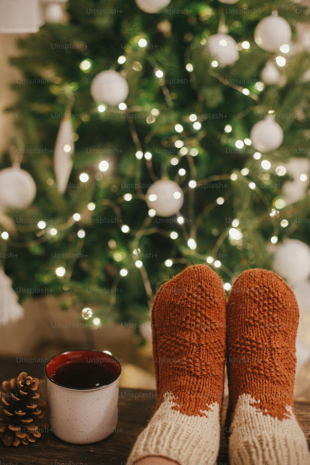 Cozy winter moments at home. Woman feet in cozy woolen socks and cup of warm tea on background of christmas tree in lights in festive evening room. Stylish warm socks on female legs