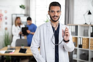 Close up horizontal shot of professional Middle Eastern male medical practitioner in white gown, demonstrating his stethoscope to camera with smile. Two doctors discussing the diagnosis on background